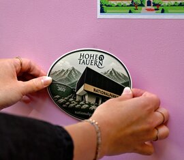 Hände bringen einen Nationalparkwelten-Sticker auf dem rosa Enzi an. | © Ferienregion Nationalpark Hohe Tauern