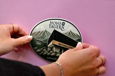 Hände bringen einen Nationalparkwelten-Sticker auf dem rosa Enzi an. | © Ferienregion Nationalpark Hohe Tauern